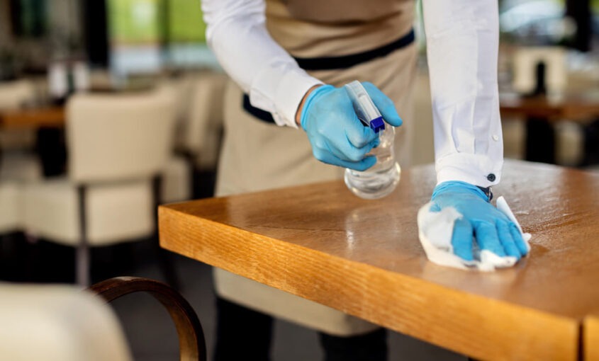Trabajador de hostelería con guantes azules desinfectando una mesa de restaurante con pulverizador y paño, siguiendo el protocolo del plan de limpieza y desinfección.